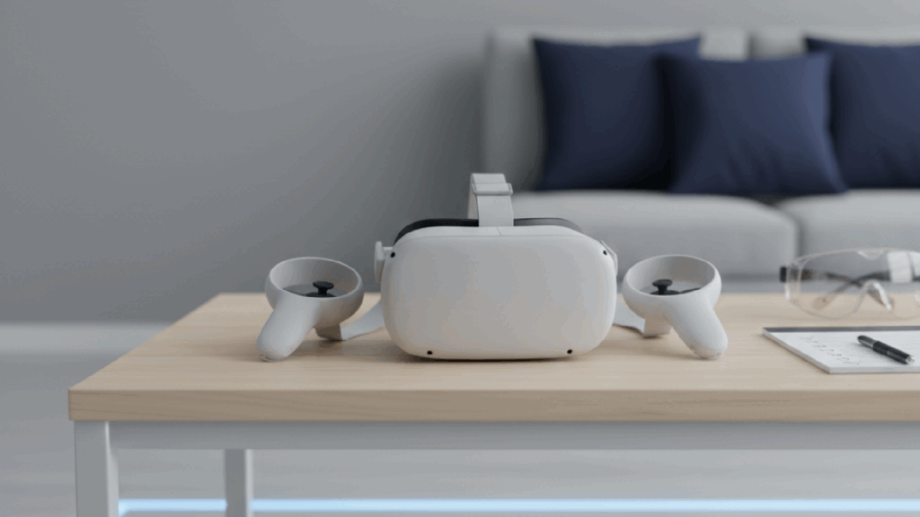 A white VR headset and two controllers sit on a light wooden coffee table in a modern living room. To the right, there is a notepad with a pen and a pair of clear safety glasses. In the blurred background, a grey sofa with dark blue cushions is visible.